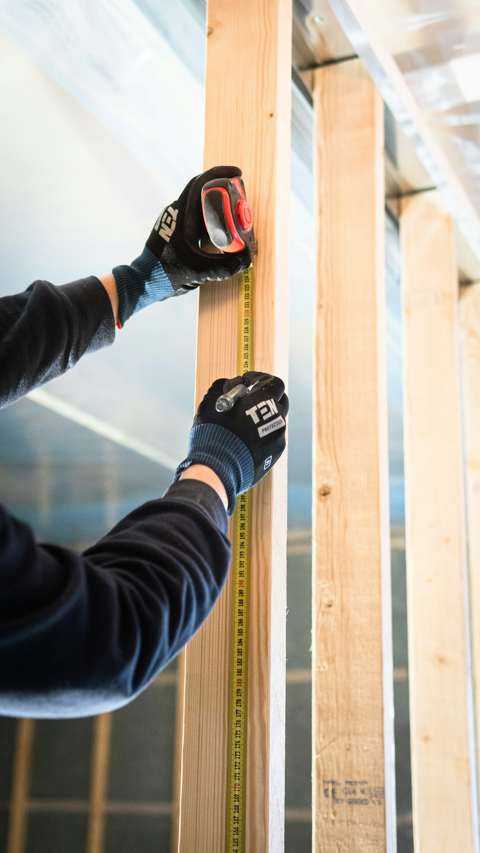 Gloved hands measuring a timber stud with a tape and marking it with a pencil