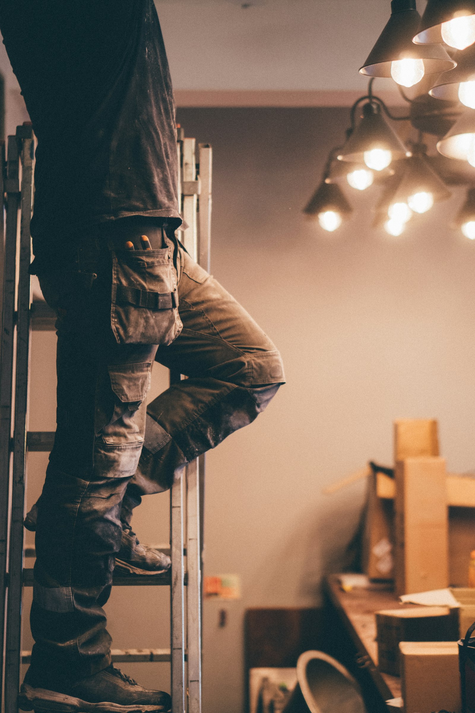 A person in work trousers standing on a ladder beneath a cluster of lights